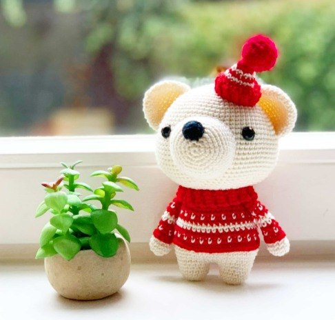 White crochet teddy bear wearing a red sweater and pom-pom hat beside a small plant